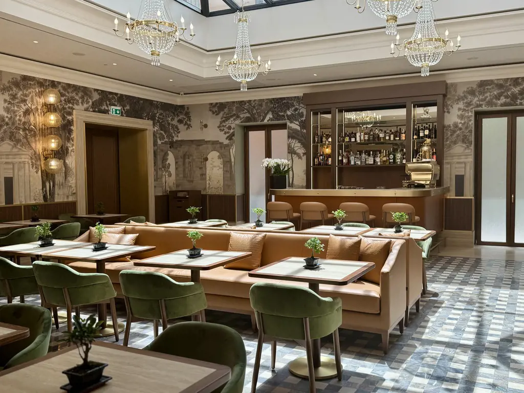 Área do Convivio Lounge no Hotel Imperiale em Roma, com sofás de couro, poltronas verdes, mesas elegantes, bar completo e lustres sob um teto de vidro.