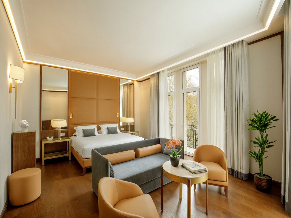 Suíte do Hotel Imperiale em Roma com cama king-size, sofá, poltronas, mesa redonda com flores, grandes janelas com cortinas claras e varanda com vista para a cidade.