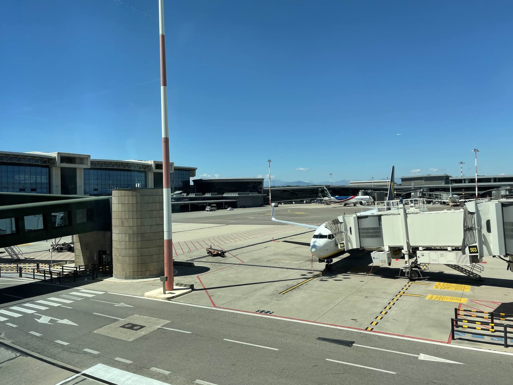 Aeroporto de Milão Malpensa fecha uma pista de março a maio de 2026 1 Aeroporto de Milão Malpensa fecha uma pista de março a maio de 2026 - Pátio de estacionamento de aeronaves no Aeroporto de Milão Malpensa com avião comercial conectado ao terminal por ponte de embarque, céu azul ao fundo