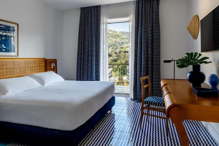 Grand Hotel Flora em Sorrento: hotel histórico reabre renovado em abril de 2026 1 Quarto do Grand Hotel Flora em Sorrento com piso em azulejo azul artesanal, cama de casal com cabeceira em madeira entalhada, escrivaninha, cortinas listradas azul e branco e varanda com vista para a vegetação