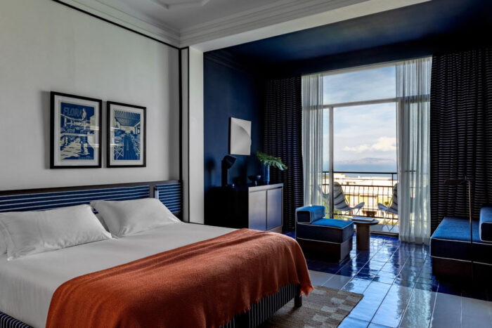 Grand Hotel Flora em Sorrento: hotel histórico reabre renovado em abril de 2026 2 Suite Junior do Grand Hotel Flora em Sorrento com parede e teto azul-marinho, cama de casal com manta terracota, piso em azulejo azul brilhante, dois quadros decorativos com a palavra Flora e varanda com vista para o mar