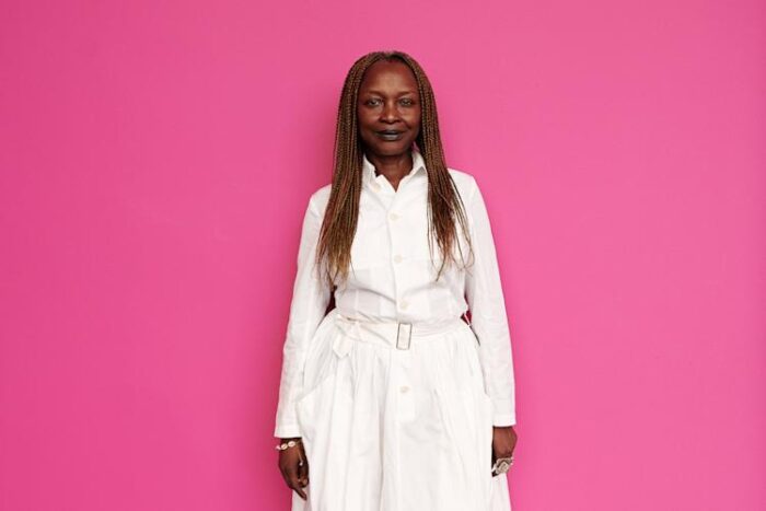 Koyo Kouoh, curadora da 61º edição da Bienal de Arte de Veneza, falecida em maio de 2025 antes da inauguração oficial. Foto da mulher africana vestida de branco com fundo rosa pink. 