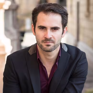 Páscoa na Sicília 2026: Música Clássica no Politeama de Palermo 2 Retrato fotográfico do maestro Giacomo Sagripanti. Um homem jovem de cabelos escuros curtos e barba aparada, vestindo um paletó preto elegante. Ele olha diretamente para a câmera com uma expressão séria e concentrada. O fundo está desfocado.