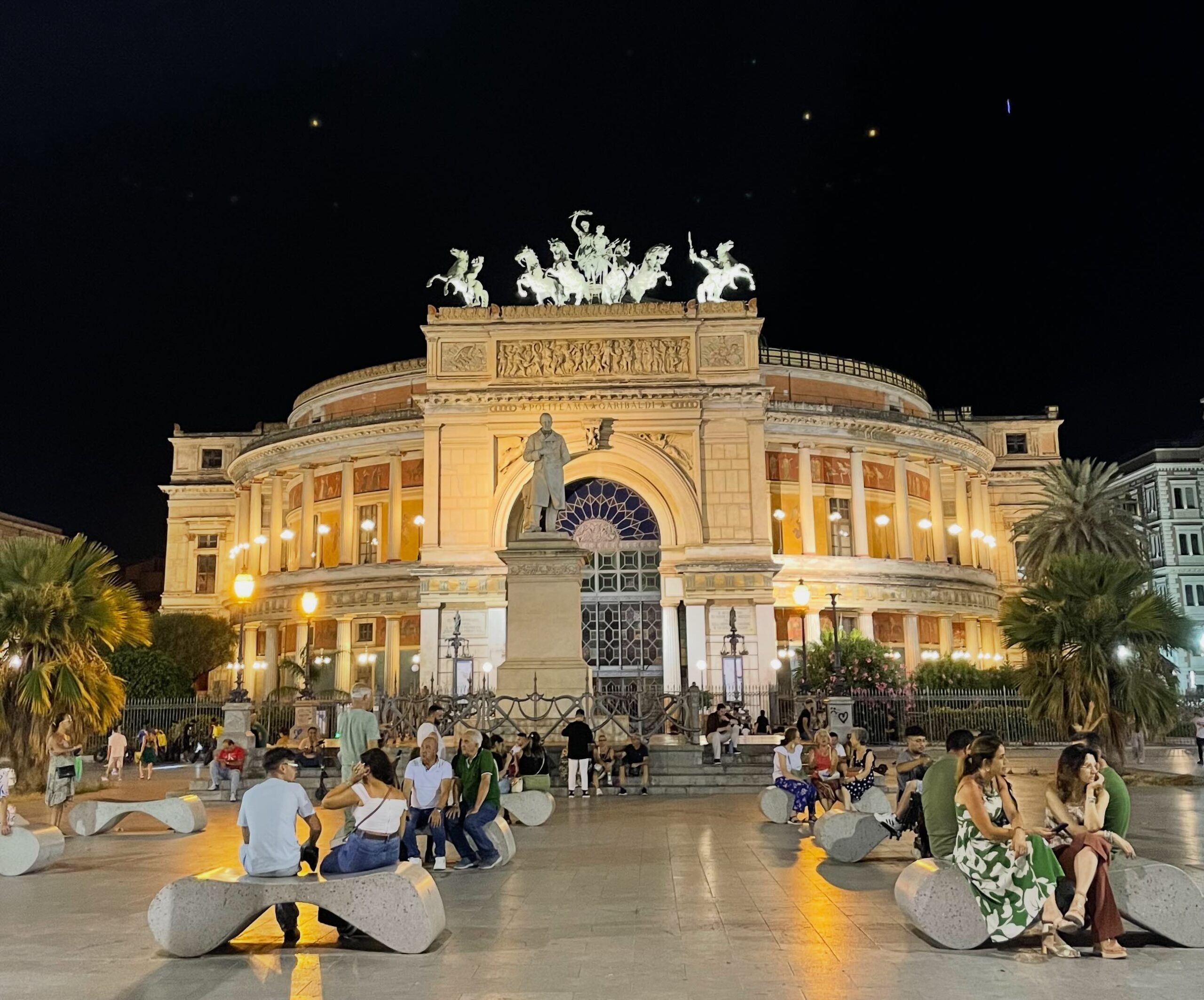 Páscoa na Sicília 2026: Música Clássica no Politeama de Palermo 1 Vista noturna da fachada do Teatro Politeama Garibaldi de Palermo iluminada por luzes quentes. Em primeiro plano, pessoas sentadas em bancos de pedra modernos na praça. Ao centro, o imponente arco do triunfo do teatro com a estátua equestre de bronze (Quadriga) no topo.