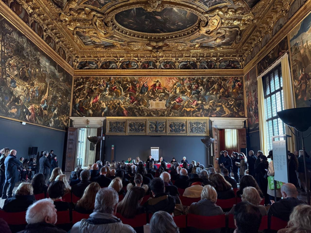 Público acompanha a apresentação da mostra “Etruschi e Veneti. Acque, culti e santuari” em uma sala ornamentada do Palazzo Ducale, em Veneza, com teto dourado e grandes pinturas históricas nas paredes.