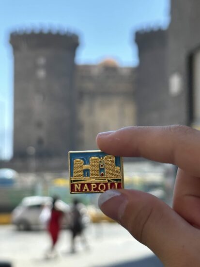A foto mostra um broche escrito Napoli com a imagem de um castelo que está como fundo da foto. Foto foi tirada em Nápoles.