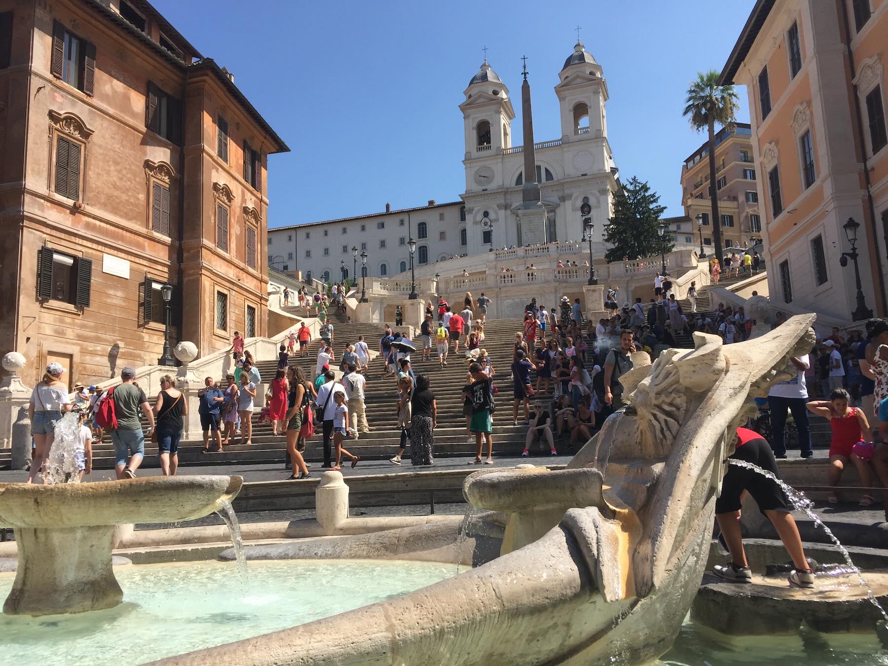Roma no verão: cuidados para montar um roteiro sem cair em roubadas 1 Fontana della Barcaccia na Piazza di Spagna, em Roma, com turistas na escadaria e a igreja Trinità dei Monti ao fundo em um dia ensolarado.